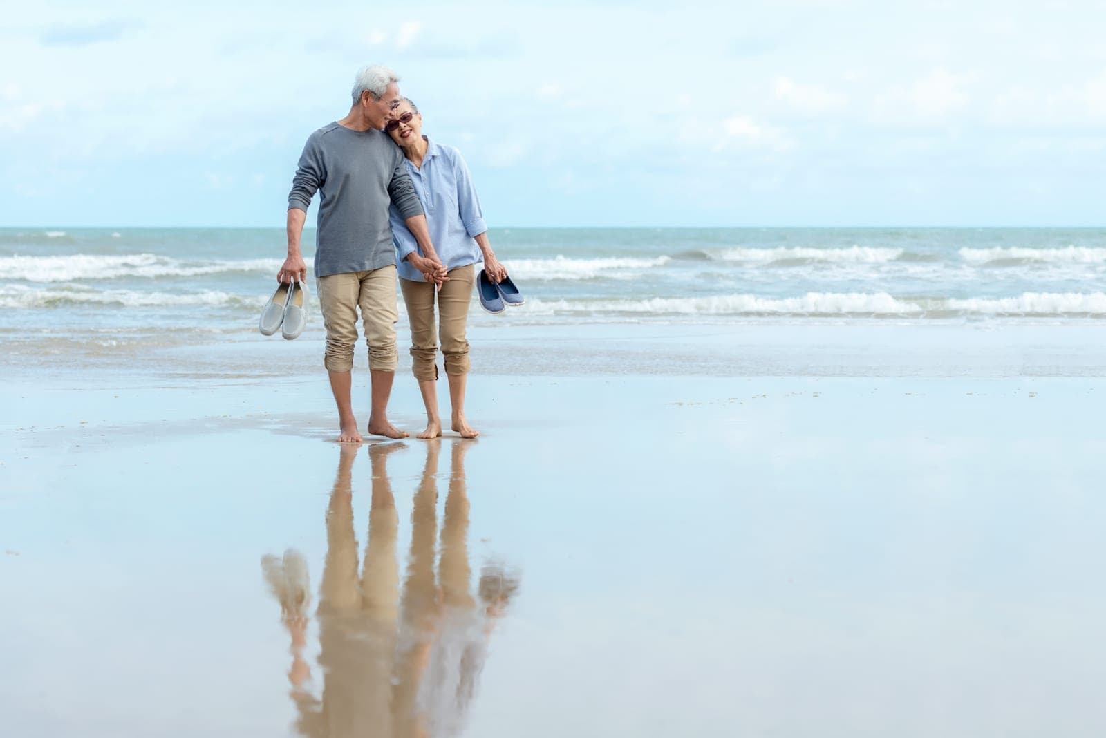 An older couple walking barefoot together along the wet sand at the water’s edge, carrying their shoes